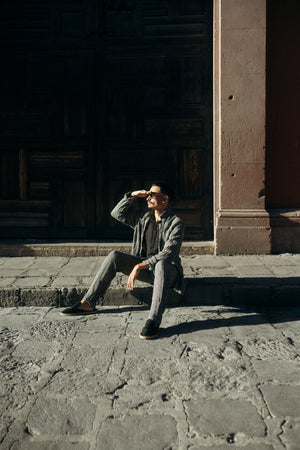 Man wearing a linen striped set, sitting on a stone step in an urban setting, shielding his eyes from the sun.