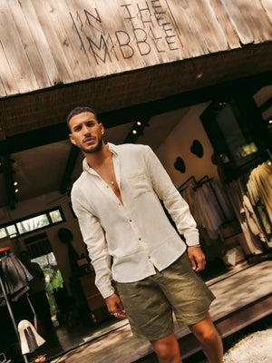Man wearing a white shirt and green shorts standing in front of a wooden building with 'In the Middle' sign.