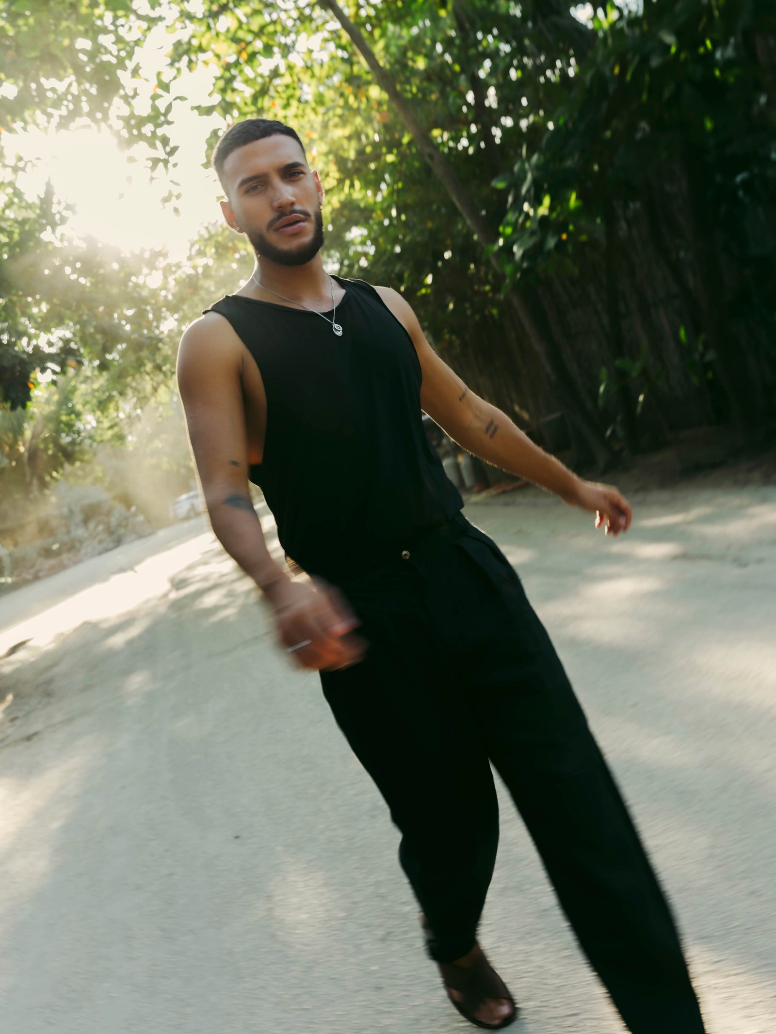 Man in black sleeveless top and tailored linen pants standing outdoors with trees in the background