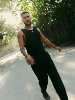 Man in black sleeveless top and tailored linen pants standing outdoors with trees in the background