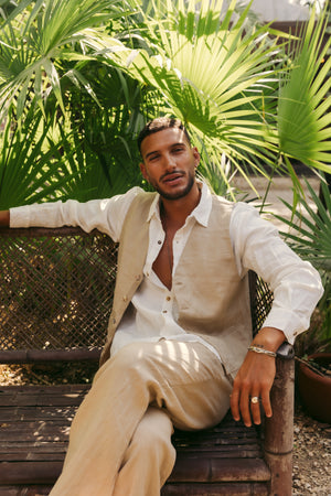 Man sitting on a wooden bench with palm leaves in the background wearing a linen set