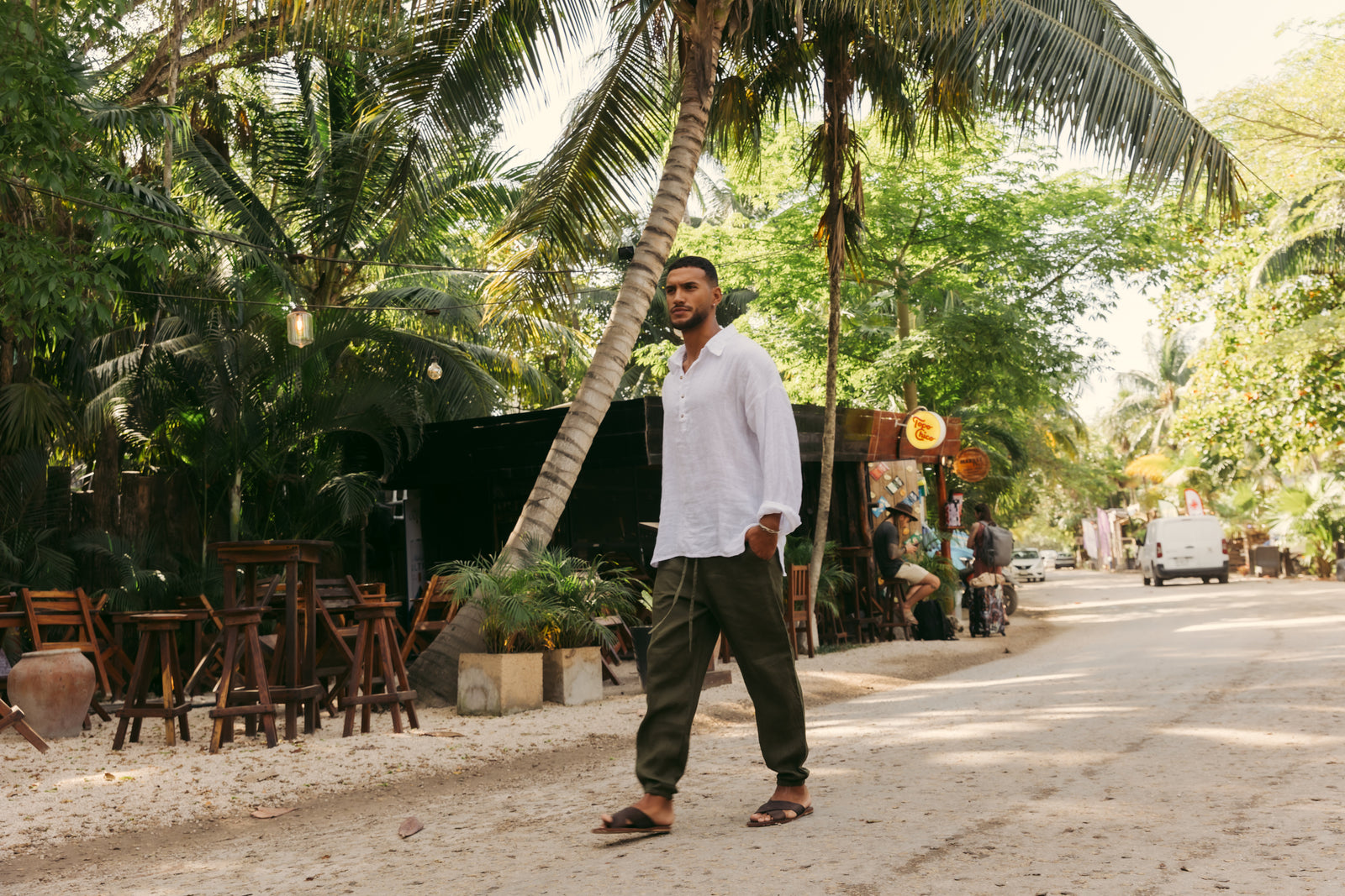 A man wearing a white long-sleeve button-up shirt, dark olive pants, and sandals walks along a sunlit dirt road lined with palm trees and outdoor seating.