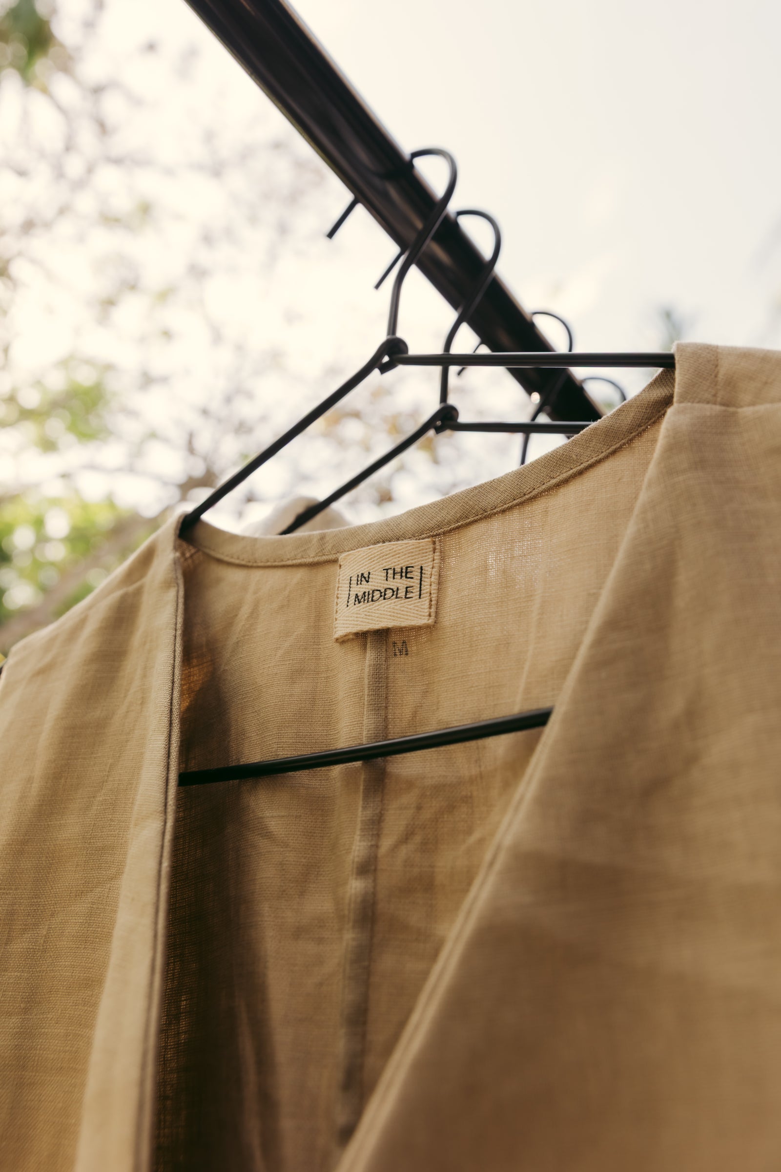 A beige linen vest or lightweight jacket hanging on a black clothes hanger outdoors. The brand label 'IN THE MIDDLE!' is visible at the collar.