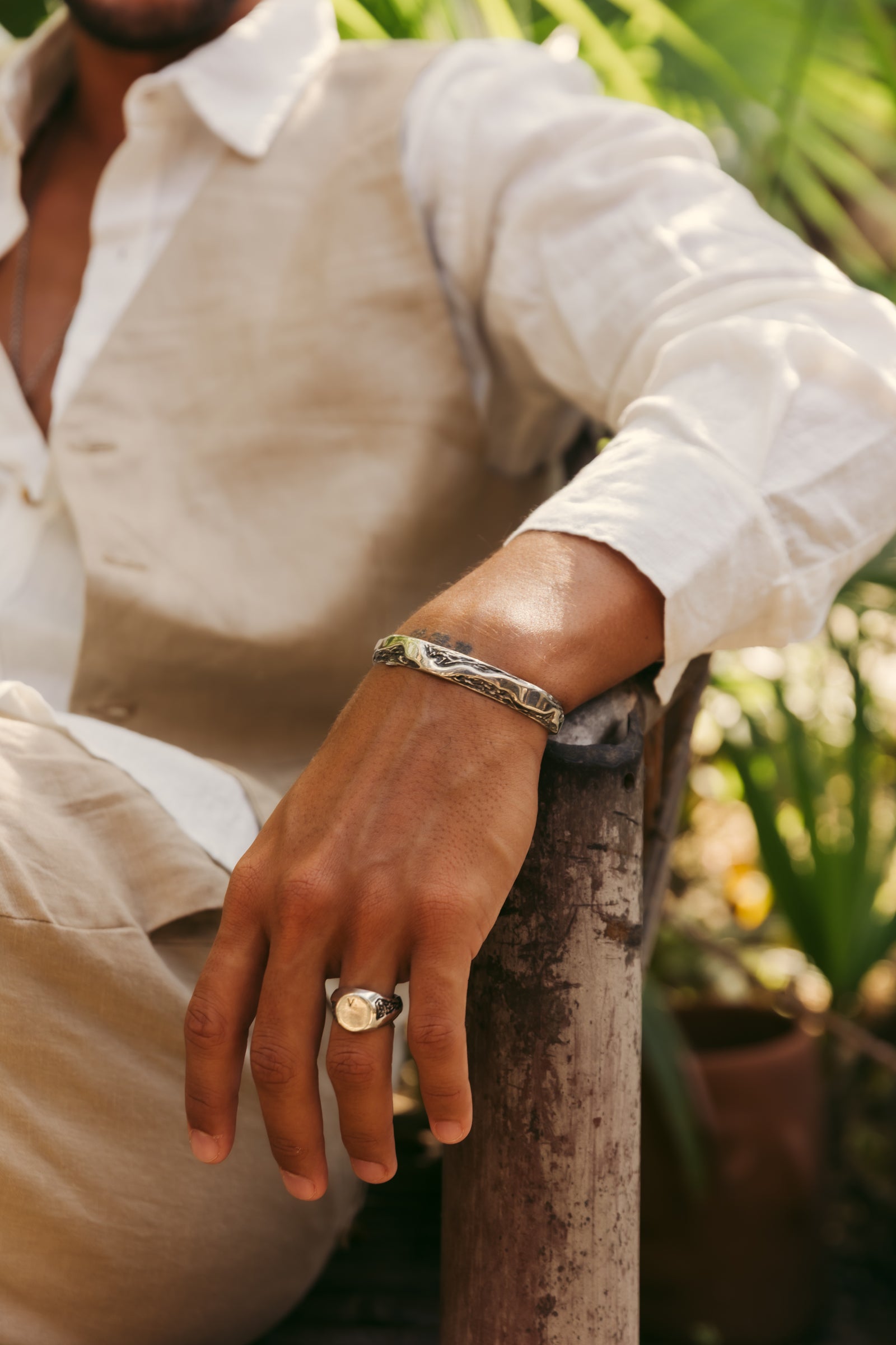 A man wearing a silver-toned snake-print cuff bracelet and a silver-toned signet ring, dressed in a white shirt and beige vest, leaning on a rustic wooden post with a blurred green garden background.