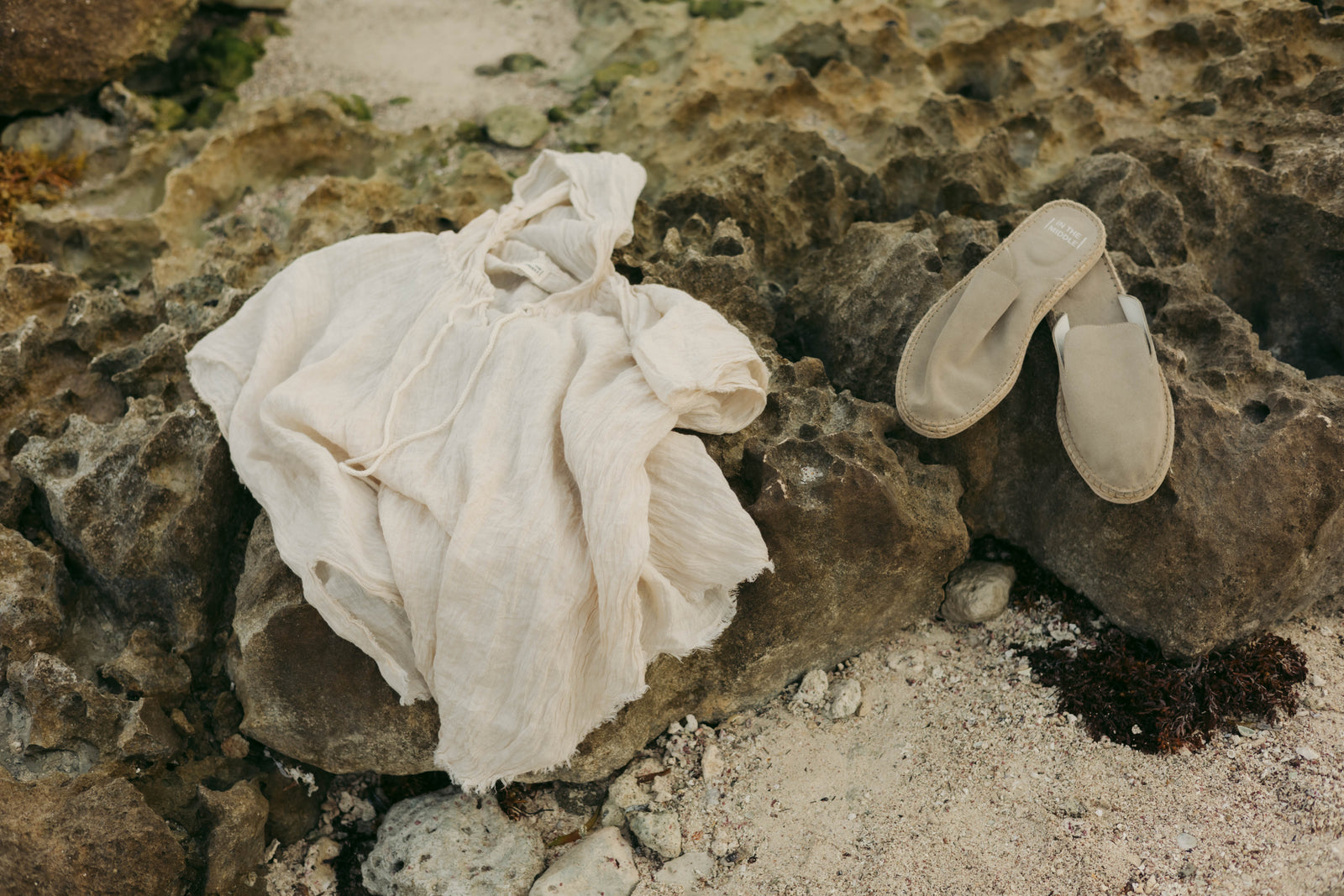 A cream-colored linen shirt and a pair of beige slip-on sandals resting on a rocky beach.