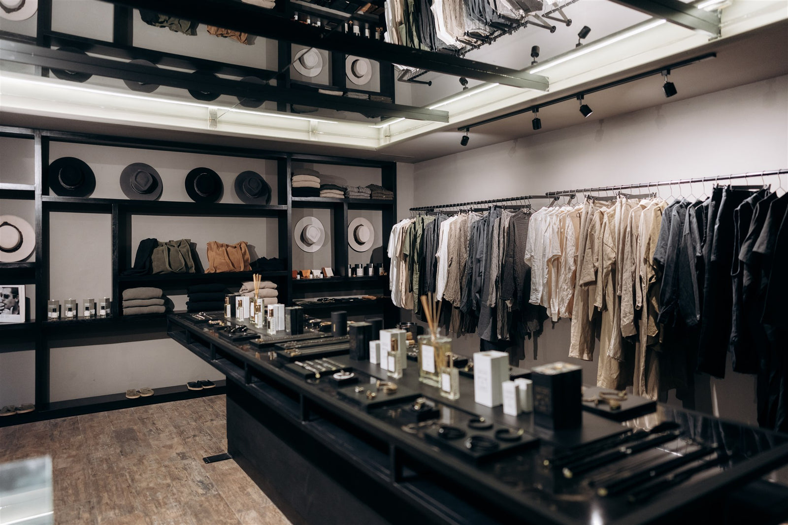 A modern men´s clothing boutique interior with racks of neutral-toned garments, shelves of hats and folded textiles, and a dark display counter in the foreground.