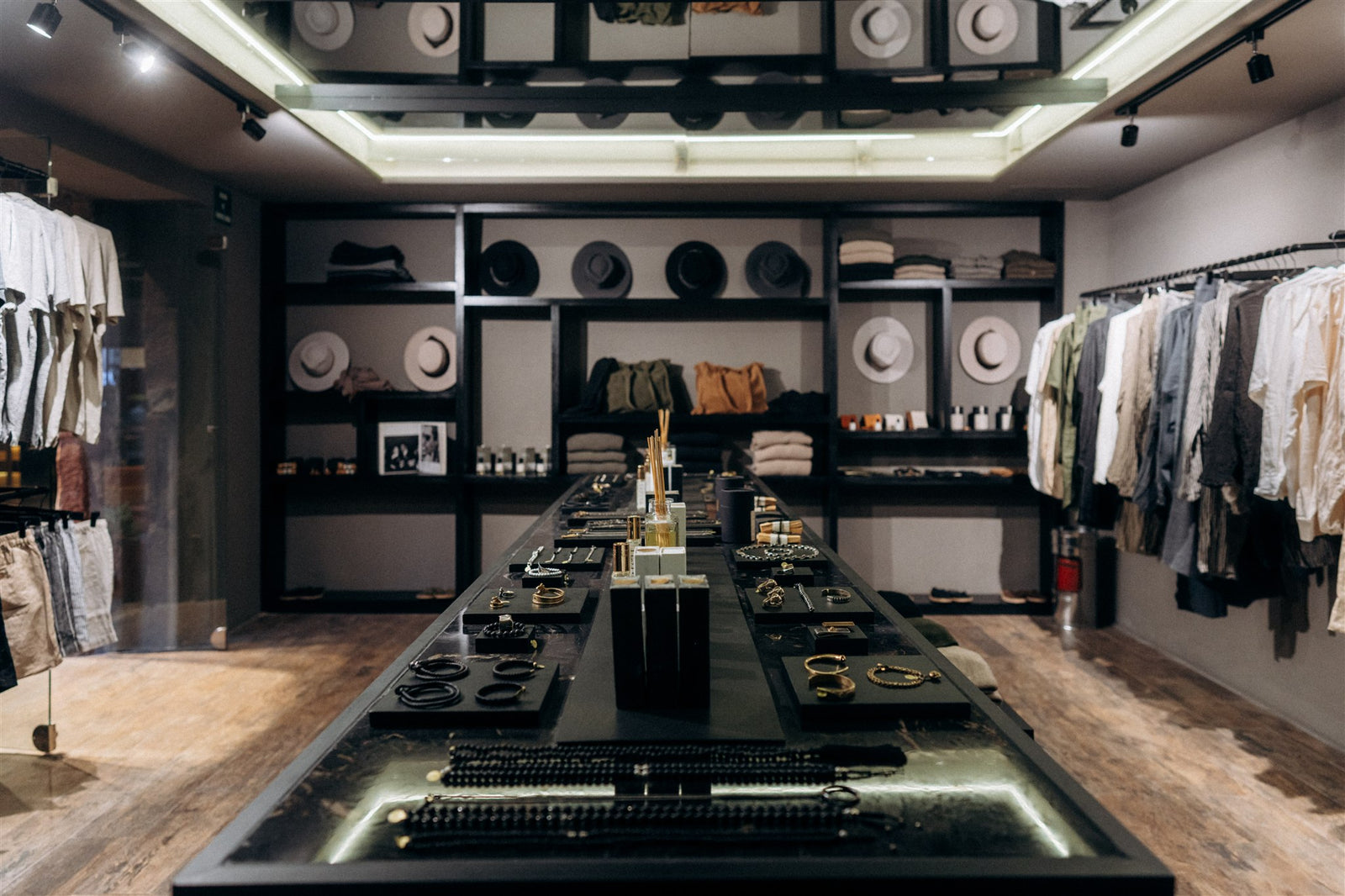 A luxury retail interior featuring a long black display table in the center with jewelry and accessories, flanked by clothing racks on both sides and styled shelves in the background.