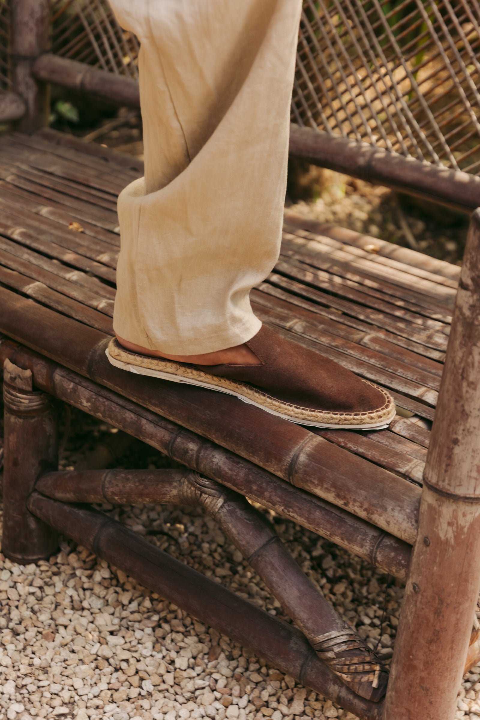 A person wearing beige trousers and a brown espadrille slip-on shoe with a jute-wrapped sole, standing on a rustic bamboo bench outdoors.