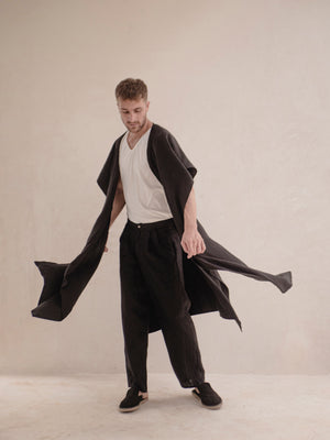 A person standing and modeling a long, black ruana with a open front, wrapped around the shoulders like a scarf.