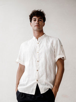 A man wearing a black linen safari style shirt with a mandarin collar and short sleeves buttoned at the cuffs. The shirt is short-sleeved and the background is neutral.