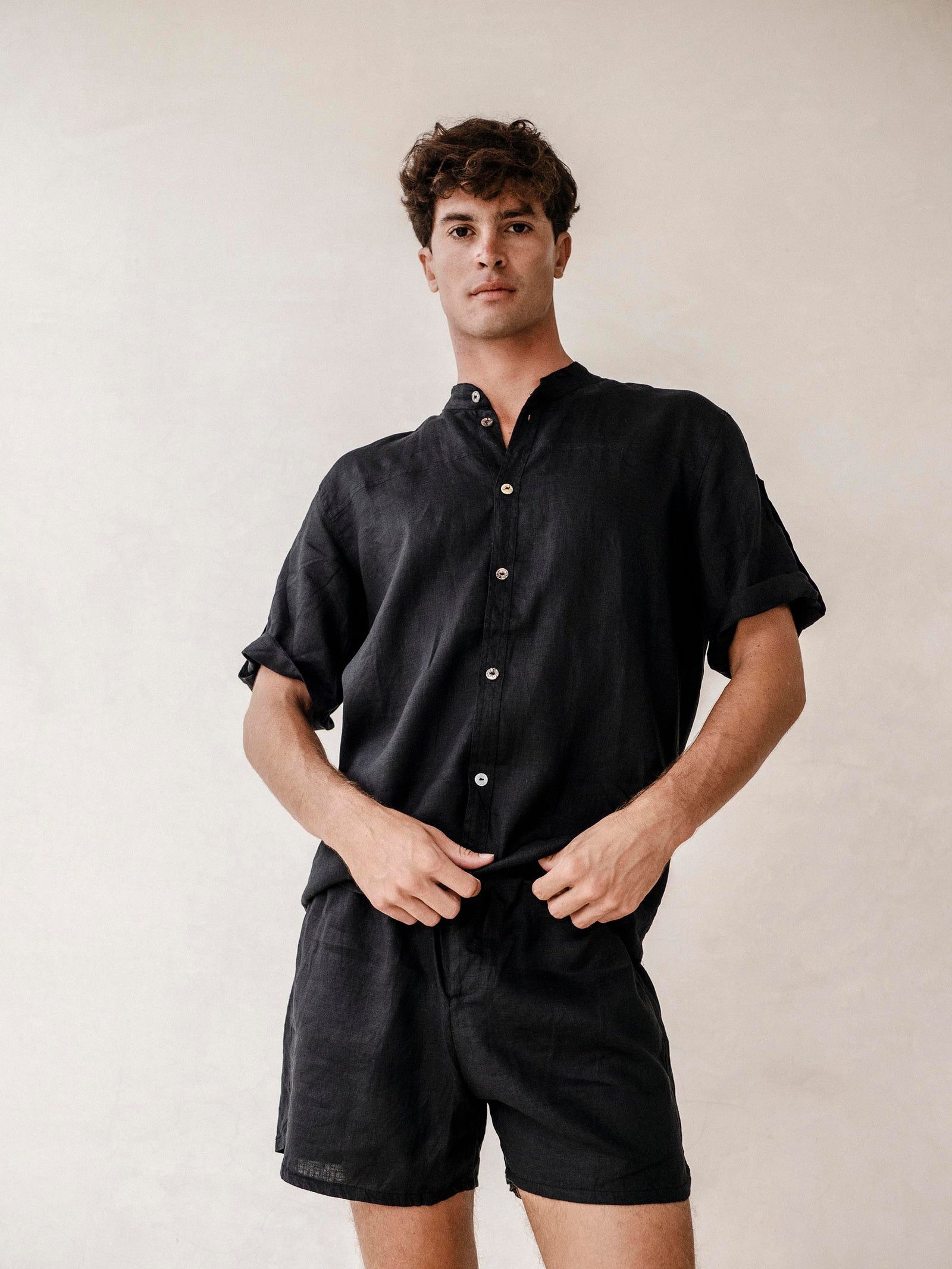 A man wearing a black linen safari style shirt with a mandarin collar and short sleeves buttoned at the cuffs. The shirt is short-sleeved and the background is neutral.