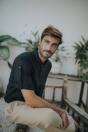 A man wearing a black linen safari style shirt with a mandarin collar and short sleeves buttoned at the cuffs. The shirt is short-sleeved 