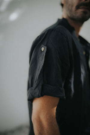 A man wearing a black linen safari style shirt with a mandarin collar and short sleeves buttoned at the cuffs. The shirt is short-sleeved and the background is neutral.