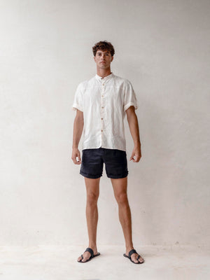 A man wearing a black linen safari style shirt with a mandarin collar and short sleeves buttoned at the cuffs. The shirt is short-sleeved and the background is neutral.
