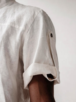 A man wearing a white linen safari style shirt with a mandarin collar and short sleeves buttoned at the cuffs. The shirt is short-sleeved and the background is neutral.