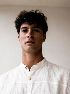 A man wearing a white linen safari style shirt with a mandarin collar and short sleeves buttoned at the cuffs. The shirt is short-sleeved and the background is neutral.