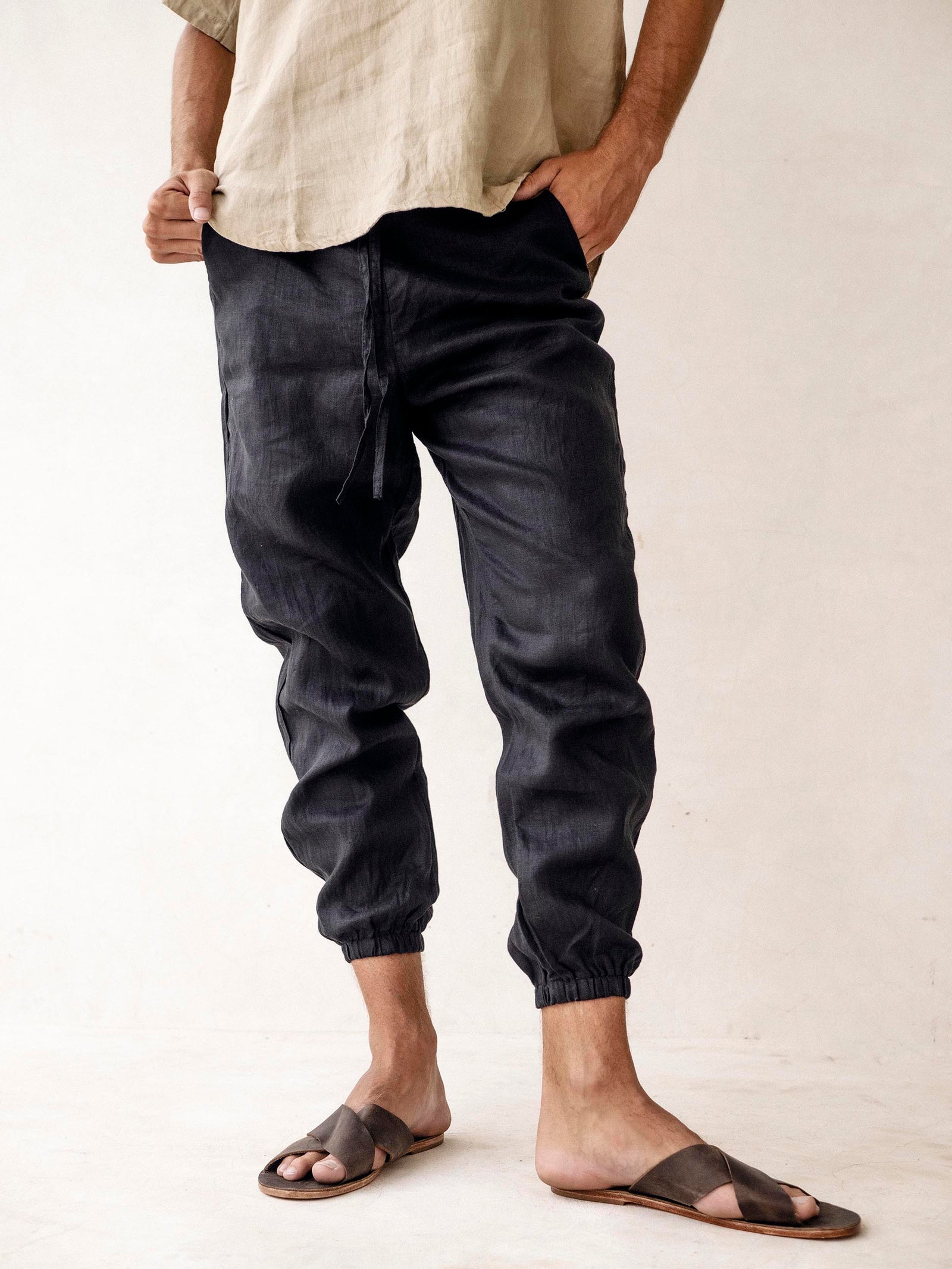 A pair of men's linen jogger pants in black, with an elasticized drawstring waist and side pockets. The pants are displayed on a model wearing sandals.