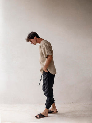 A pair of men's linen jogger pants in black, with an elasticized drawstring waist and side pockets. The pants are displayed on a model wearing sandals.