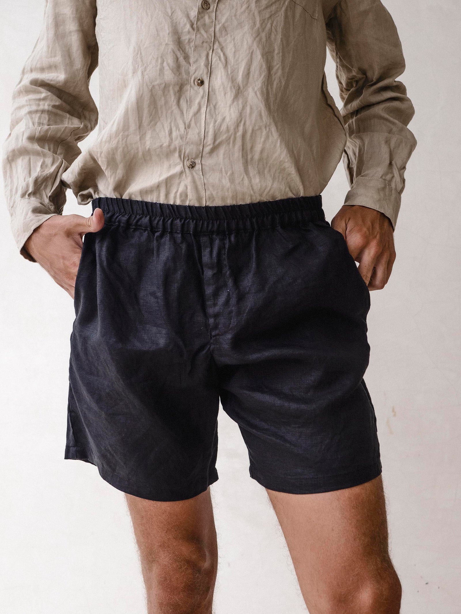 A man wearing black linen bermuda shorts with elasticized waist and side pockets. He is also wearing a light-colored shirt.