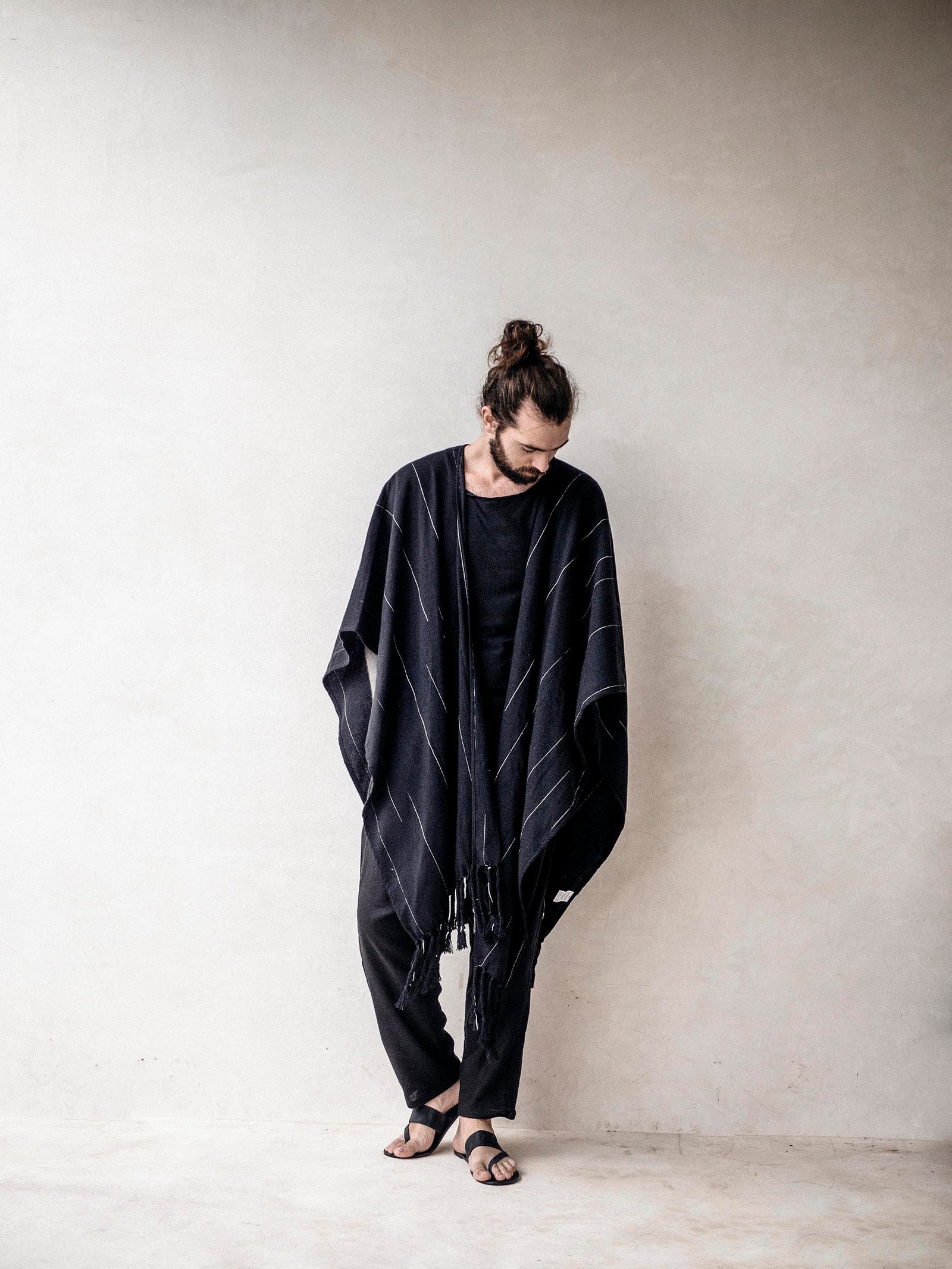 A man wearing a black cape poncho with fringes, standing in a minimalistic setting against a white background.