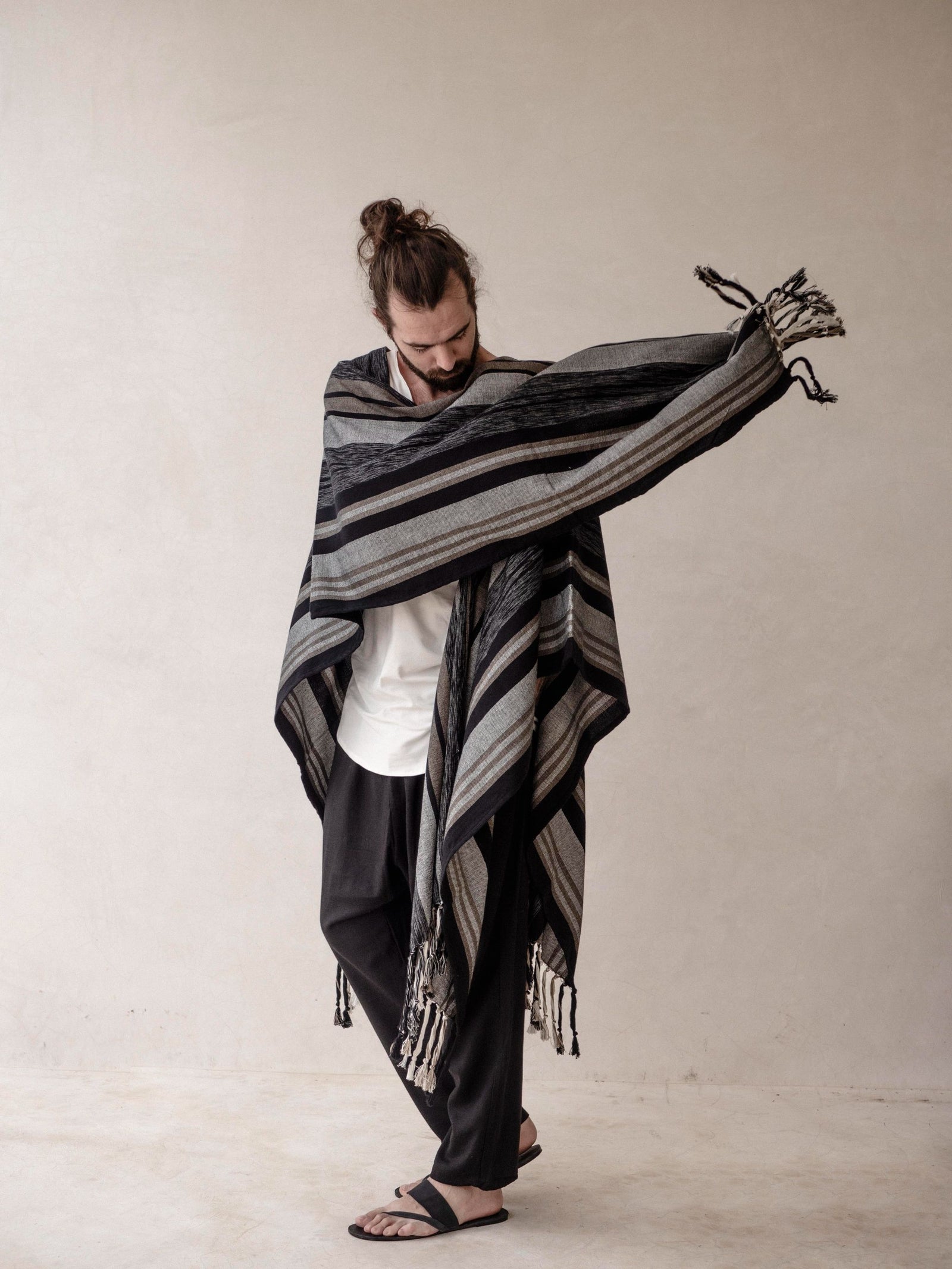A man wearing a striped cape poncho with fringes standing in front of a neutral background.