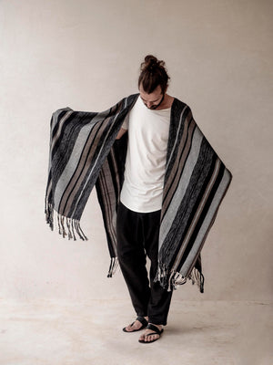 A man wearing a striped cape poncho with fringes standing in front of a neutral background.