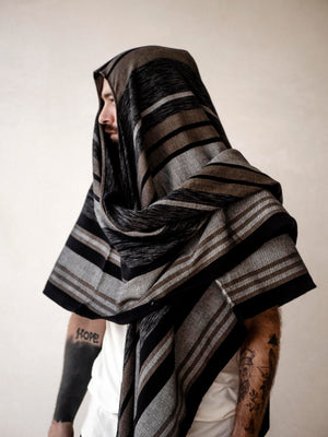A man wearing a striped cape poncho with fringes standing in front of a neutral background.