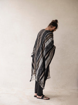 A man wearing a striped cape poncho with fringes standing in front of a neutral background.