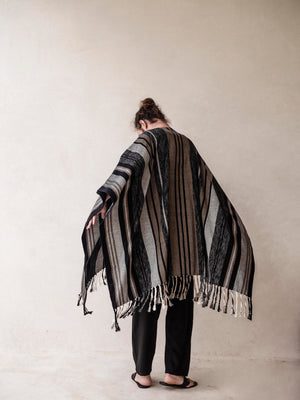 A man wearing a striped cape poncho with fringes standing in front of a neutral background.