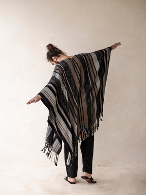 A man wearing a striped cape poncho with fringes standing in front of a neutral background.