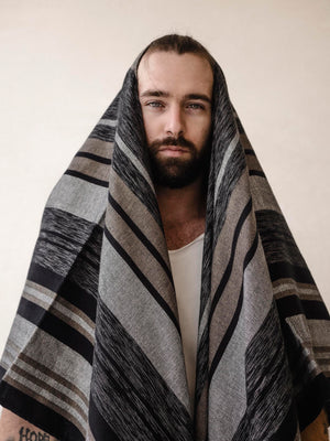 A man wearing a striped cape poncho with fringes standing in front of a neutral background.