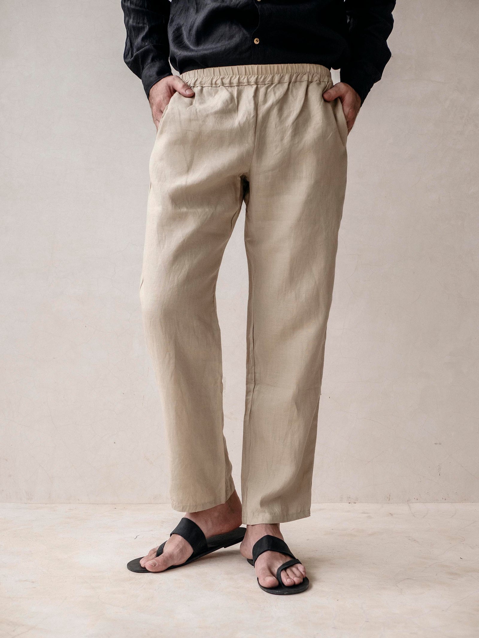 A man standing with hands in pockets, wearing beige linen pants and a dark top.
